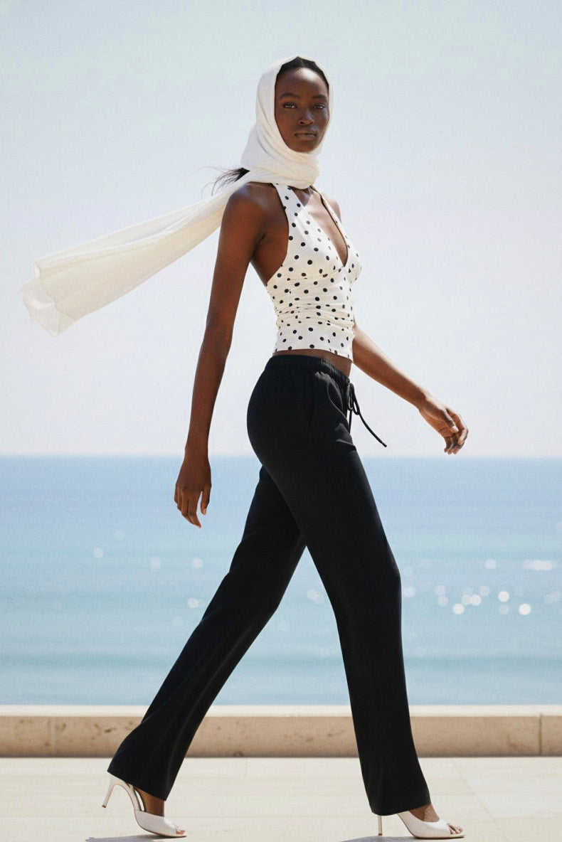 Woman walking by the ocean wearing a white polka dot top and black pants with a beige scarf.