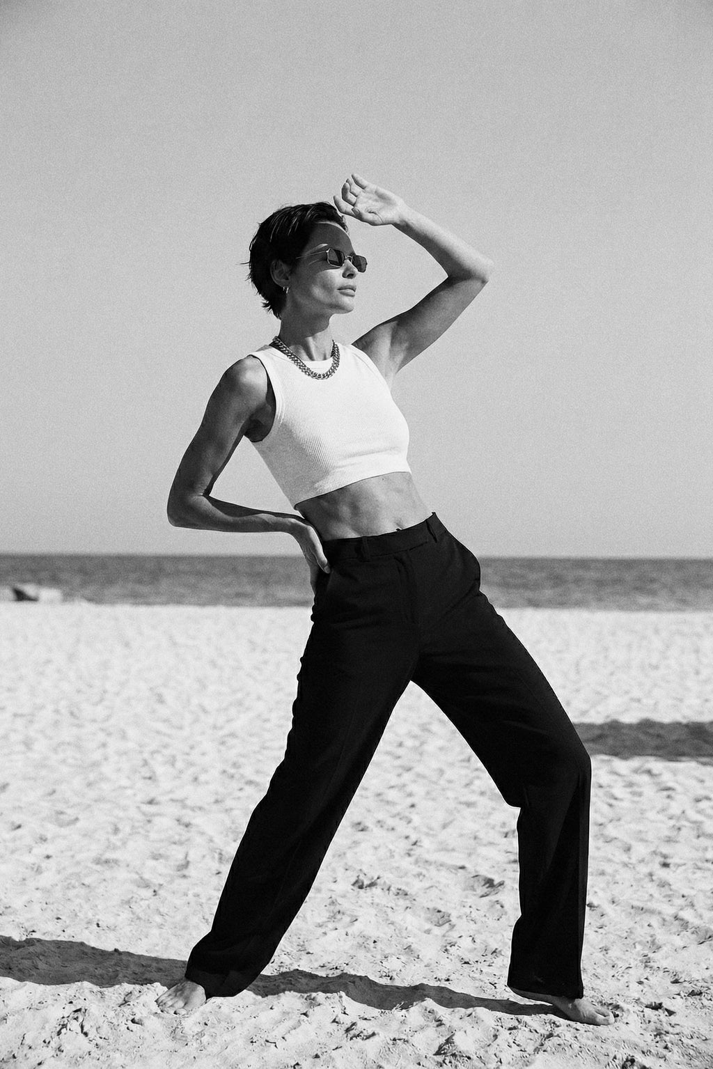 Person posing on a sandy beach wearing a white top and black pants.