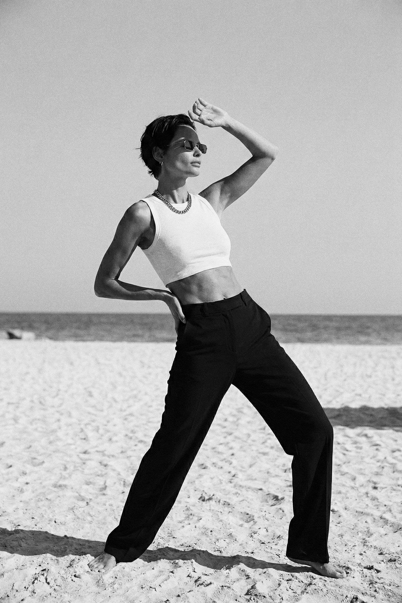 Person posing on a sandy beach wearing a white top and black pants.