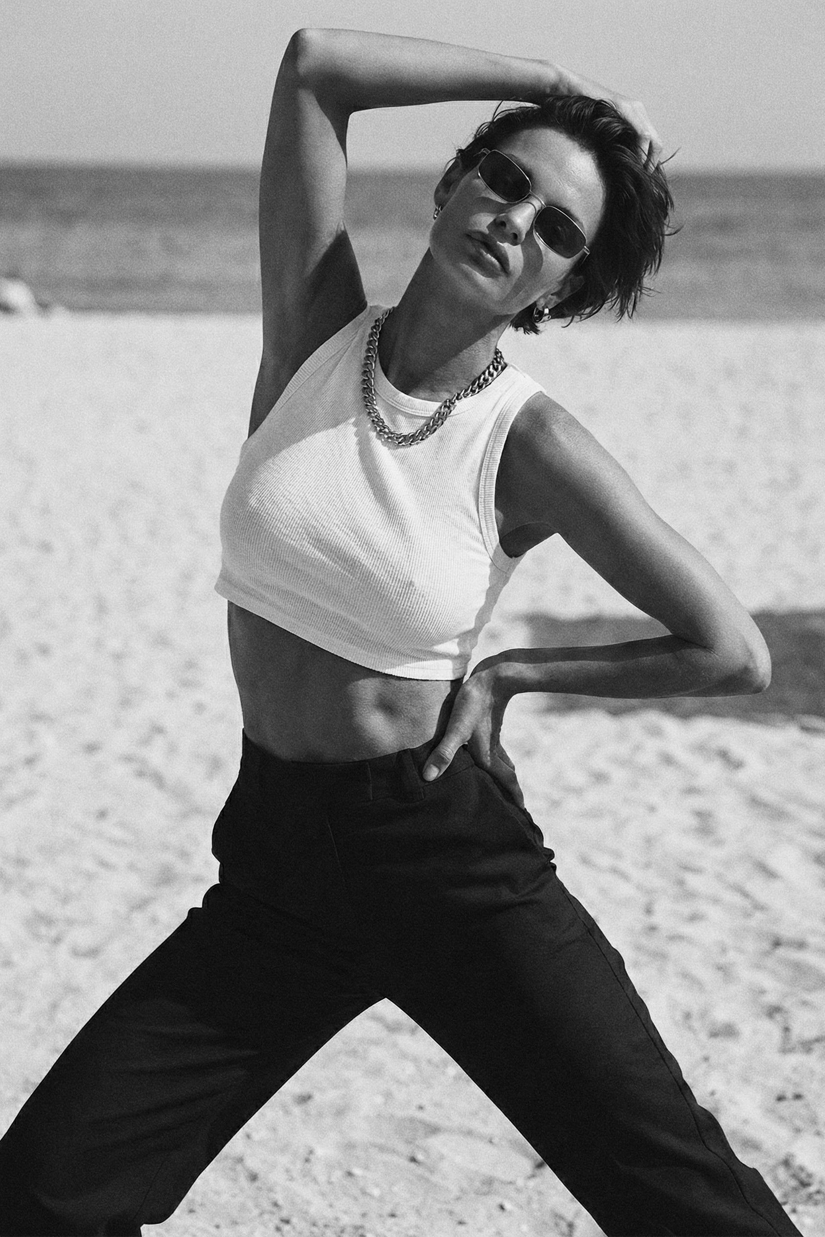 Person posing on a beach wearing a white sleeveless top and black pants.