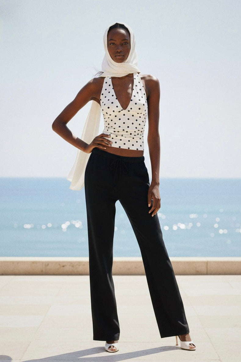 Woman wearing a white sleeveless top with black polka dots and black pants, standing against a clear blue sky.