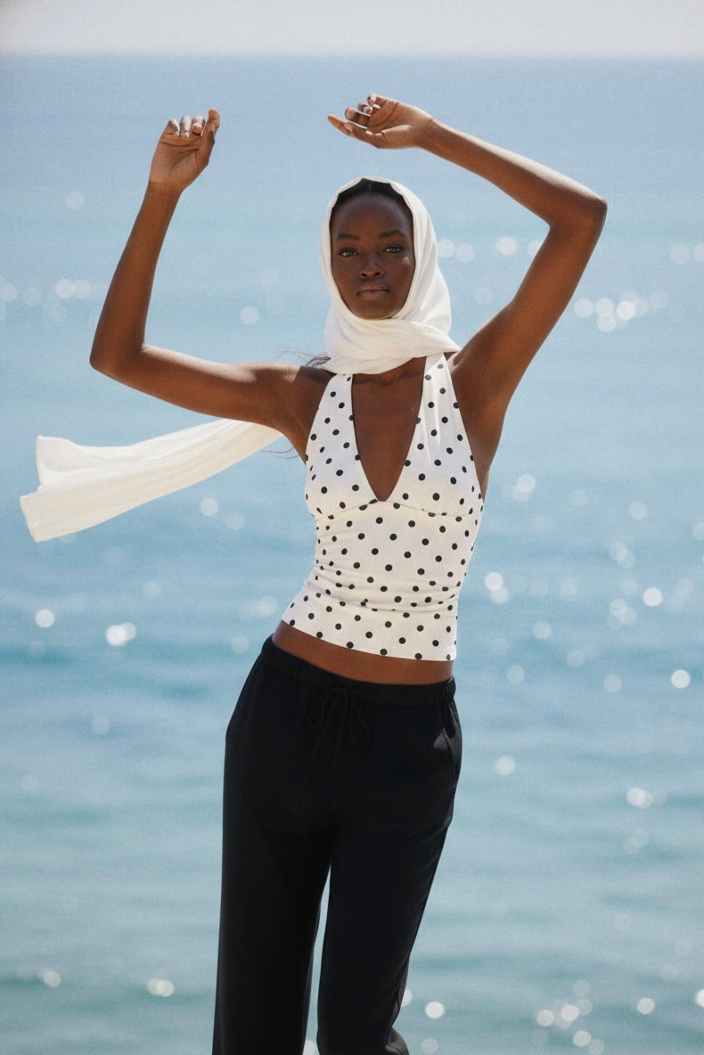 Woman in a polka dot top and black pants with a white scarf, standing against a blue water background.
