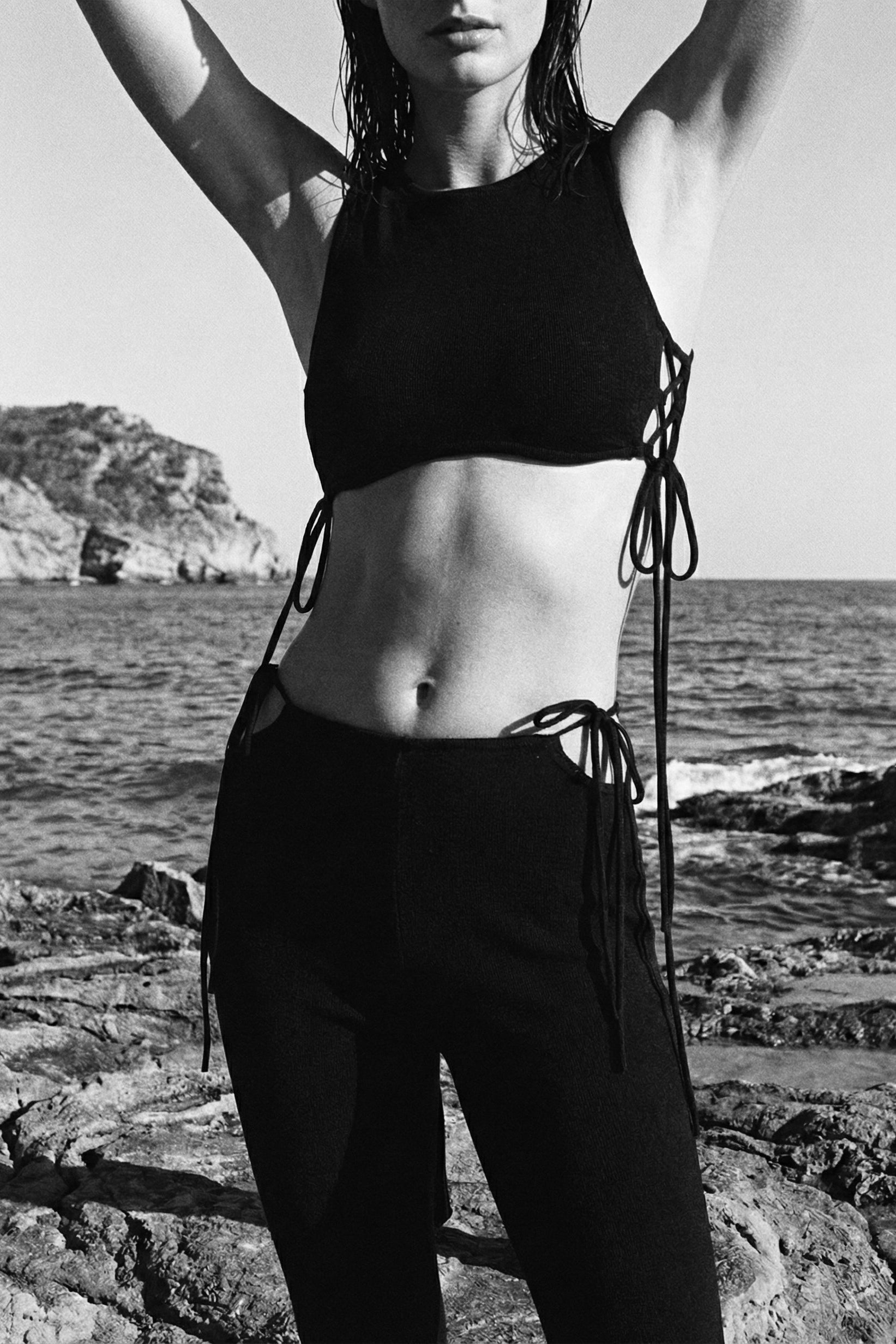 Person wearing a black bikini top and pants standing on rocky terrain by the sea.