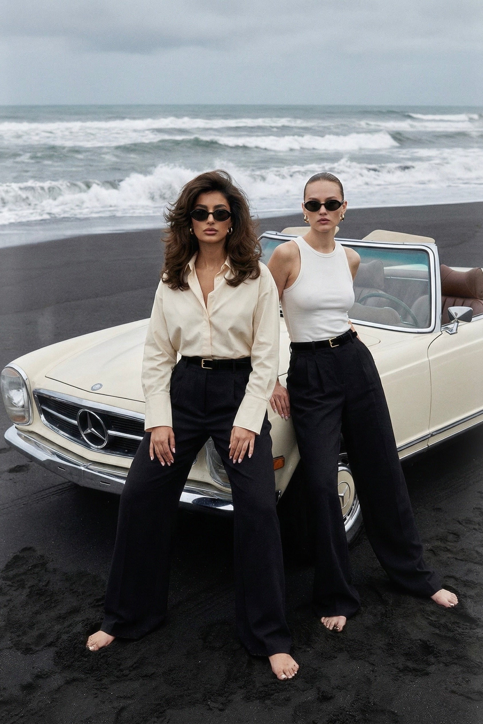 Two women standing in front of a vintage car on a beach with ocean waves in the background.